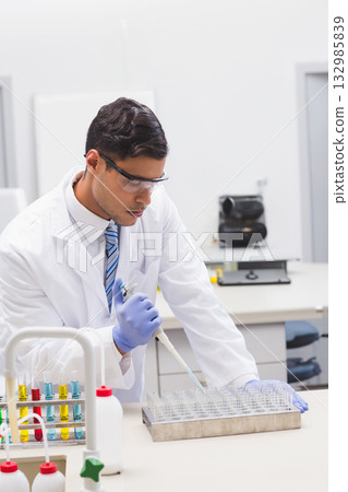 Asian man wearing lab coat using micropipette on lab bench dispensing liquid into multi-well plate 132985839
