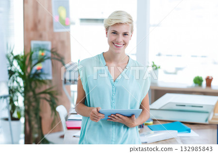 Woman smiling and holding tablet in bright modern office with documents, folders, scanner Woman smiling and holding tablet in bright modern office with documents, folders, scanner 132985843