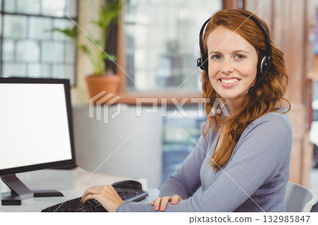 Female office worker wearing headset using keyboard and mouse at desk near window with monitor 132985847