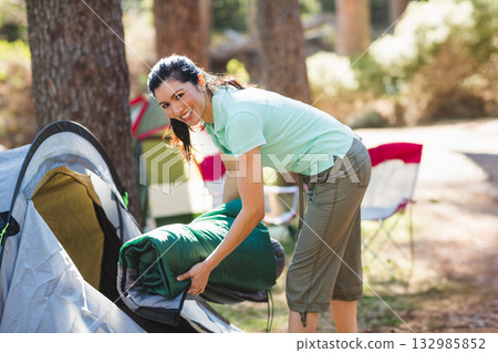 Female camper placing green sleeping bag into partially pitched tent near folding chairs in forest Female camper placing green sleeping bag into partially pitched tent near folding chairs in forest 132985852
