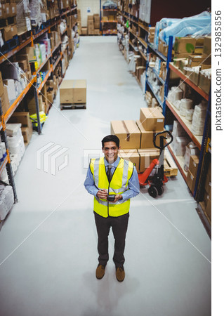 Male warehouse worker in safety vest scanning boxes in warehouse aisle with scanner and pallet jack 132985856