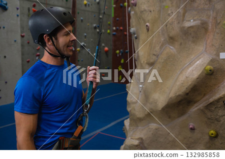 Mid adult man standing at rock-climbing gym wearing helmet and harness holding belay rope 132985858