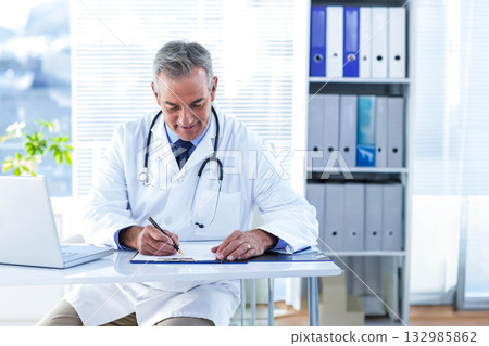 Senior male doctor sitting at desk writing on clipboard wearing stethoscope in medical office Senior male doctor sitting at desk writing on clipboard wearing stethoscope in medical office 132985862