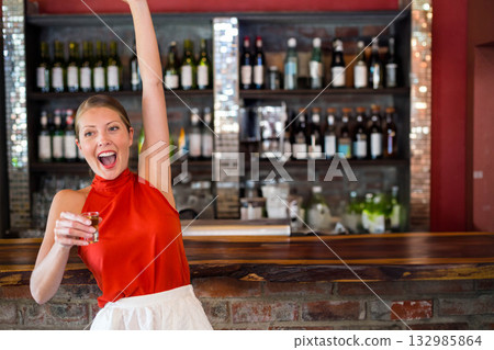 Mid adult woman raising arm holding shot glass among liquor bottles at bar counter, copy space Mid adult woman raising arm holding shot glass among liquor bottles at bar counter, copy space 132985864