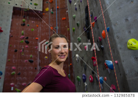 Woman in twenties wearing burgundy tee smiling against climbing wall with multicolored holds, ropes 132985878