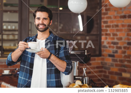 Barista man wearing blue shirt at counter holding white cup saucer near espresso machine Barista man wearing blue shirt at counter holding white cup saucer near espresso machine 132985885