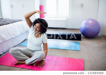 African American woman performing yoga side stretch on pink mat beside exercise ball in bedroom 132985889