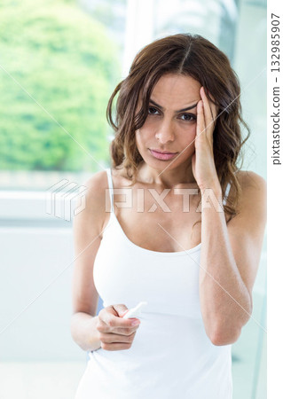 Middle-aged woman holding slim plastic test device and touching temple in bright bathroom by window 132985907