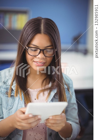 Teenage student wearing eyeglasses, denim jacket sitting at desk in classroom holding white tablet 132985911