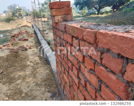 Boundary wall under construction using red bricks and cement mortar at rural site, showing traditional bricklaying process and step-by-step layering of bricks for compound wall building Boundary wall under construction using red bricks and cement mortar at rural site, showing traditional bricklaying process and step-by-step layering of bricks for compound wall building 132986250