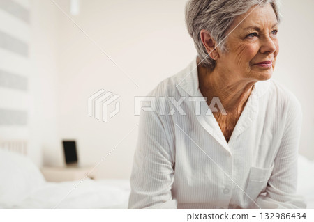 Senior woman sitting on bed in bedroom wearing pajamas looking at device on nightstand, copy space 132986434
