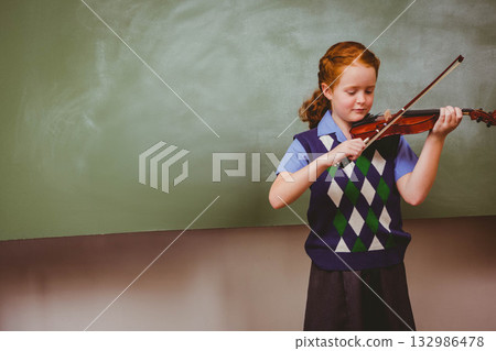 Schoolgirl playing violin with bow before green chalkboard in classroom wearing uniform, copy space 132986478
