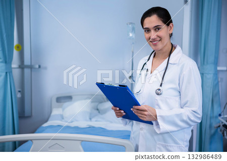 Smiling Indian doctor holding clipboard, wearing stethoscope at bedside with IV pole, copy space Smiling Indian doctor holding clipboard, wearing stethoscope at bedside with IV pole, copy space 132986489