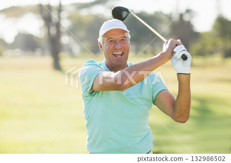 Senior man swinging driver and smiling finishing golf swing on fairway in polo shirt and glove Senior man swinging driver and smiling finishing golf swing on fairway in polo shirt and glove 132986502