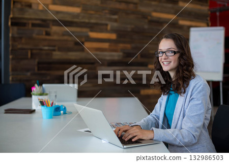 Woman in teal blouse, glasses typing on laptop at coworking table with notebook, copy space Woman in teal blouse, glasses typing on laptop at coworking table with notebook, copy space 132986503