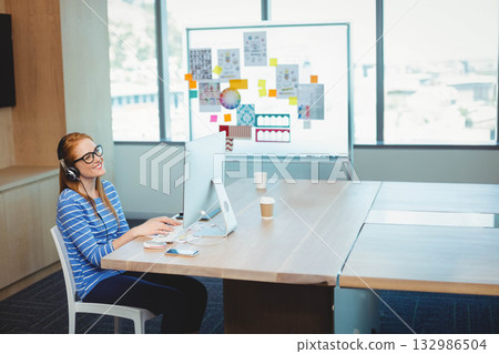 Woman wearing headset working on monitor at wooden table in office with coffee cups, copy space Woman wearing headset working on monitor at wooden table in office with coffee cups, copy space 132986504