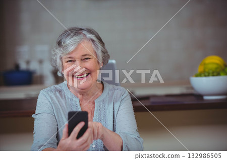 Senior woman tapping smartphone while sitting at countertop in kitchen near laptop and fruit bowl Senior woman tapping smartphone while sitting at countertop in kitchen near laptop and fruit bowl 132986505