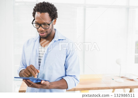 African American man standing by window blinds in office tapping blue tablet screen, copy space 132986511
