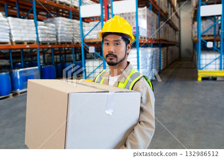 Asian male warehouse worker wearing safety vest holding cardboard box near racks in warehouse 132986512