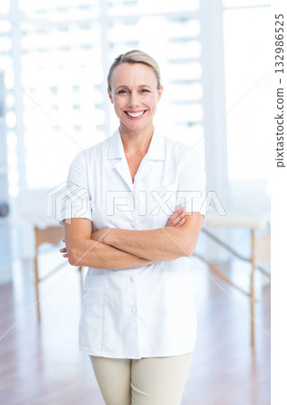 Female therapist smiling with crossed arms wearing tunic in clinic by massage table under blinds 132986525