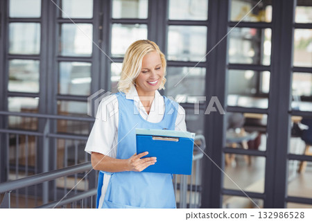 Female nurse wearing blue uniform standing in clinic corridor holding blue clipboard with papers Female nurse wearing blue uniform standing in clinic corridor holding blue clipboard with papers 132986528