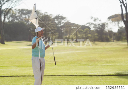 Senior man standing in golf attire on putting green holding white flagpole and club, copy space 132986531