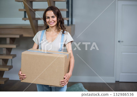 Woman carrying taped cardboard box in living room near wooden staircase and cushion Woman carrying taped cardboard box in living room near wooden staircase and cushion 132986536