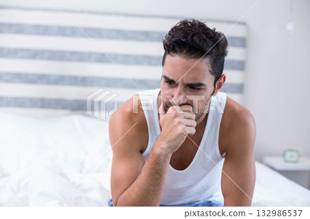 Man wearing white tank top sitting on bed edge covering mouth in bedroom with alarm clock 132986537