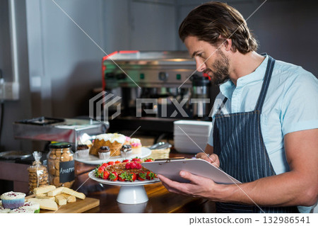 Male baker wearing apron holding clipboard examining pastries behind bakery counter, copy space Male baker wearing apron holding clipboard examining pastries behind bakery counter, copy space 132986541