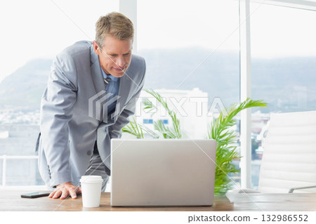 Man in grey suit leaning on desk by window in office, checking laptop screen with smartphone Man in grey suit leaning on desk by window in office, checking laptop screen with smartphone 132986552