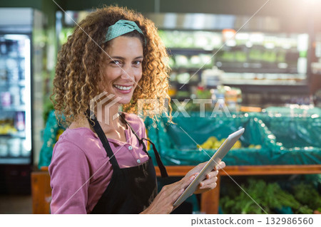 Female store employee wearing apron standing in grocery produce aisle using tablet by fruit display Female store employee wearing apron standing in grocery produce aisle using tablet by fruit display 132986560