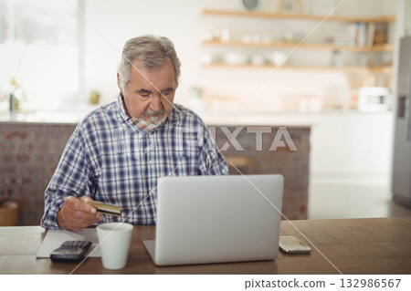Senior man sitting in kitchen entering credit card info on laptop with mug calculator and notebook 132986567