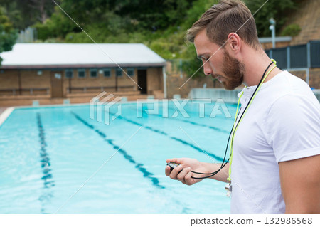Male coach holding stopwatch and checking display standing poolside with lane ropes, copy space Male coach holding stopwatch and checking display standing poolside with lane ropes, copy space 132986568