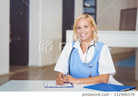 Mid adult female nurse smiling while writing on clipboard at reception desk in clinic, copy space Mid adult female nurse smiling while writing on clipboard at reception desk in clinic, copy space 132986576