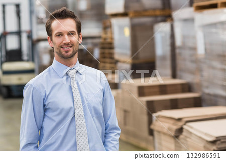Man in thirties wearing blue shirt and tie standing by forklift, pallet stacks in warehouse aisle 132986591