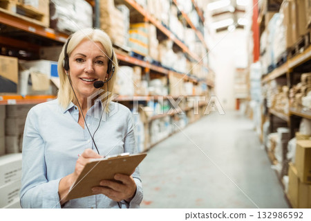 Woman checking inventory on clipboard while wearing headset among metal racks in warehouse aisle 132986592