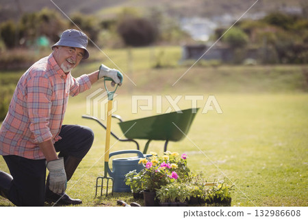 Senior man kneeling on backyard lawn wearing bucket hat, using fork beside wheelbarrow, copy space 132986608