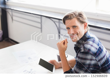 man smiling, holding tablet at white desk by window with coffee cup and papers, copy space man smiling, holding tablet at white desk by window with coffee cup and papers, copy space 132986609