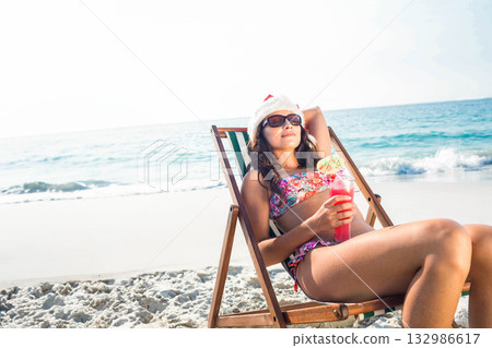 Woman in holiday hat reclining on wooden chair at water's edge holding pink cocktail, copy space 132986617