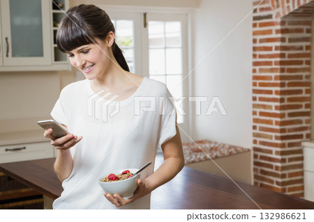 Woman standing at kitchen island, smiling at smartphone and holding cereal bowl with strawberries 132986621