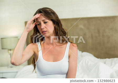 Woman holding hand to forehead and sitting on white sheets in bedroom with nightstand lamp Woman holding hand to forehead and sitting on white sheets in bedroom with nightstand lamp 132986623