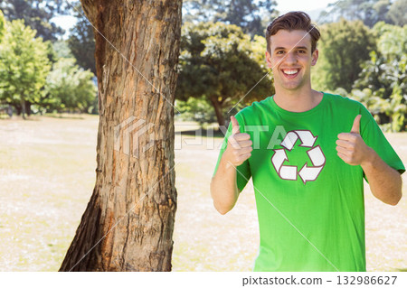 Man standing beside tree trunk in park wearing green recycling T-shirt giving thumbs up, copy space 132986627