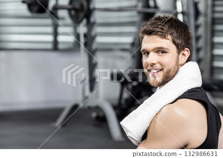 Man sitting on bench in gym wearing sleeveless top holding towel by squat rack, copy space 132986631
