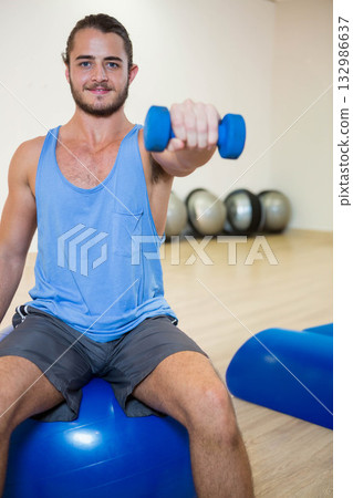 Fit man holding blue dumbbell while sitting on stability ball in gym studio wearing sportswear 132986637