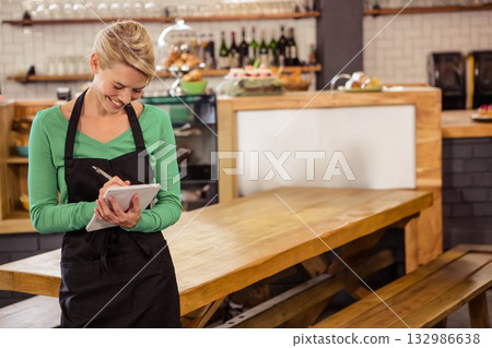 Female server in black apron writing on notepad at bakery café with pastry domes, copy space 132986638
