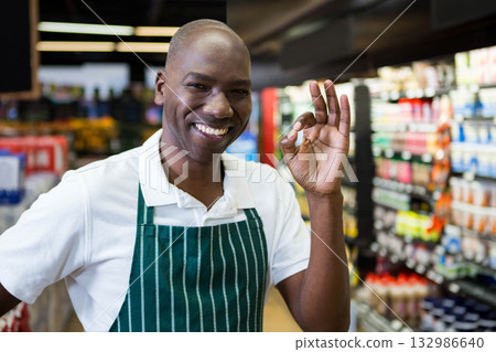 African American man wearing apron giving OK gesture while stocking store aisle with bottled drinks 132986640