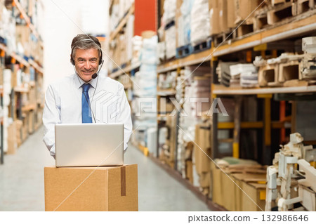 Warehouse manager operating open laptop on cardboard box in warehouse aisle with headset microphone Warehouse manager operating open laptop on cardboard box in warehouse aisle with headset microphone 132986646
