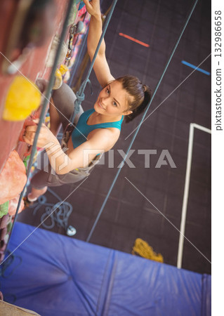 Female climber ascending artificial wall in climbing gym using rope, harness and holds, copy space Female climber ascending artificial wall in climbing gym using rope, harness and holds, copy space 132986658