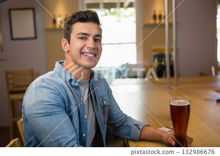 Man sitting at pub counter wearing chambray shirt sipping beer from pint glass with smartphone 132986676
