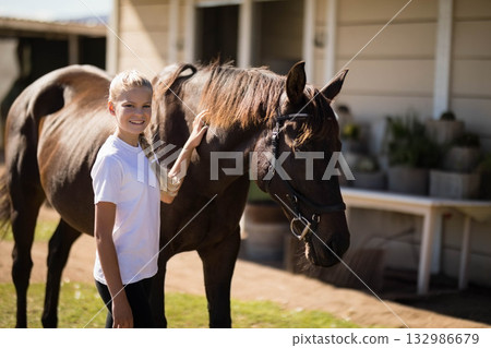 Child girl petting bridled horse in stable yard near porch with wooden table and potted plants 132986679